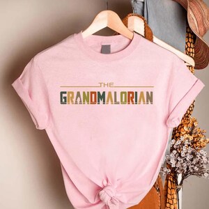 Puede incluir: Camiseta rosa claro con el texto "THE GRANDMALORIAN" en una fuente retro. El texto es multicolor, con verde, naranja y marr&oacute;n. La camiseta est&aacute; colgada en una percha de madera.