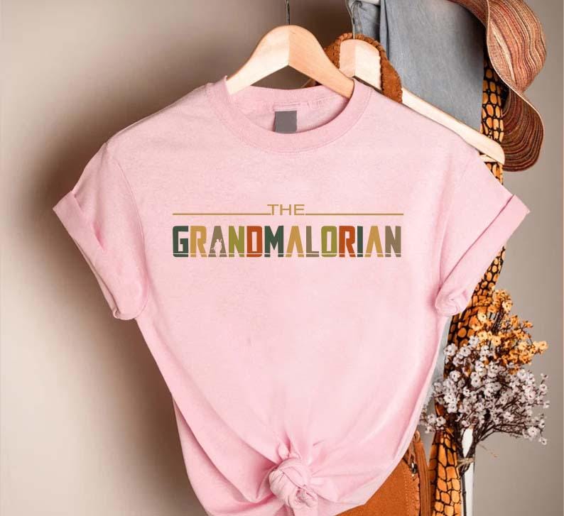 Puede incluir: Camiseta rosa claro con el texto "THE GRANDMALORIAN" en una fuente retro. El texto es multicolor, con verde, naranja y marr&oacute;n. La camiseta est&aacute; colgada en una percha de madera.