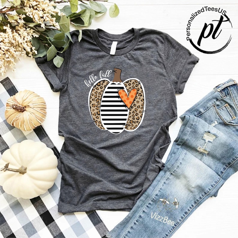 Peut inclure: T-shirt gris fonc&eacute; avec un motif de citrouille compos&eacute; d'imprim&eacute; l&eacute;opard, de rayures noires et blanches et d'un c&oelig;ur orange. Les mots "hello fall" sont &eacute;crits au-dessus de la citrouille. Le t-shirt est pr&eacute;sent&eacute; avec un jean bleu.