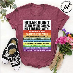 May include: Burgundy t-shirt with the text "HITLER DIDN'T START WITH CAMPS HE STARTED WITH:" followed by a list of actions in rainbow colors. Black and white sneakers are on the left. A bouquet of flowers is on the right.