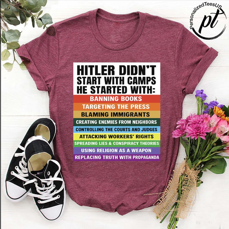 May include: Burgundy t-shirt with the text "HITLER DIDN'T START WITH CAMPS HE STARTED WITH:" followed by a list of actions in rainbow colors. Black and white sneakers are on the left. A bouquet of flowers is on the right.