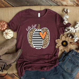 Peut inclure: T-shirt bordeaux avec un motif de citrouille &agrave; rayures noires et blanches, imprim&eacute; l&eacute;opard et un c&oelig;ur orange. Les mots "hello fall" sont &eacute;crits au-dessus de la citrouille. Le t-shirt est pr&eacute;sent&eacute; avec des d&eacute;corations d'automne et un jean.