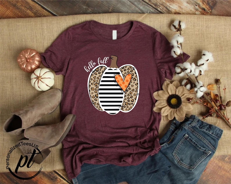 Peut inclure: T-shirt bordeaux avec un motif de citrouille &agrave; rayures noires et blanches, imprim&eacute; l&eacute;opard et un c&oelig;ur orange. Les mots "hello fall" sont &eacute;crits au-dessus de la citrouille. Le t-shirt est pr&eacute;sent&eacute; avec des d&eacute;corations d'automne et un jean.