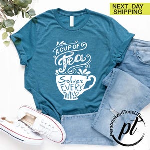 May include: Teal t-shirt with the text "A Cup of Tea Solves Everything" in white script. The design features a teacup graphic. The shirt is paired with blue jeans and white sneakers.