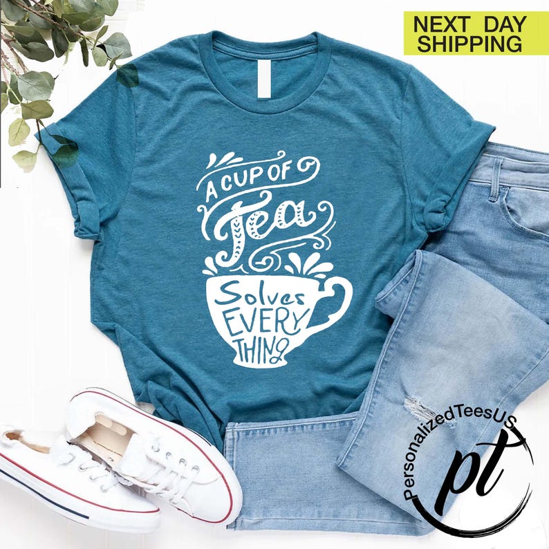 May include: Teal t-shirt with the text "A Cup of Tea Solves Everything" in white script. The design features a teacup graphic. The shirt is paired with blue jeans and white sneakers.