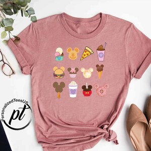 May include: A pink t-shirt with a graphic of various Disney food items, including Mickey Mouse shaped ice cream, pizza, a hamburger, and a donut. The graphic is in a cartoon style and features a variety of colors.