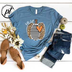 Peut inclure: Un t-shirt bleu clair avec un motif de citrouille combinant imprim&eacute; l&eacute;opard, rayures noires et blanches et un c&oelig;ur orange. Les mots "hello fall" sont &eacute;crits en cursive. Le t-shirt est associ&eacute; &agrave; un jean bleu et des chaussures marron.