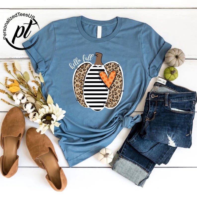 Peut inclure: Un t-shirt bleu clair avec un motif de citrouille combinant imprim&eacute; l&eacute;opard, rayures noires et blanches et un c&oelig;ur orange. Les mots "hello fall" sont &eacute;crits en cursive. Le t-shirt est associ&eacute; &agrave; un jean bleu et des chaussures marron.