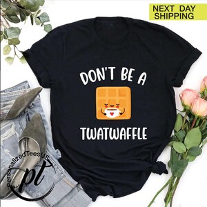 K&ouml;nnte beinhalten: Schwarzes T-Shirt mit dem wei&szlig;en Text "DON'T BE A TWATWAFFLE". Das Design zeigt eine l&auml;chelnde Waffel-Figur, die eine Kaffeetasse h&auml;lt. Das Shirt liegt auf Jeans und Schuhen.
