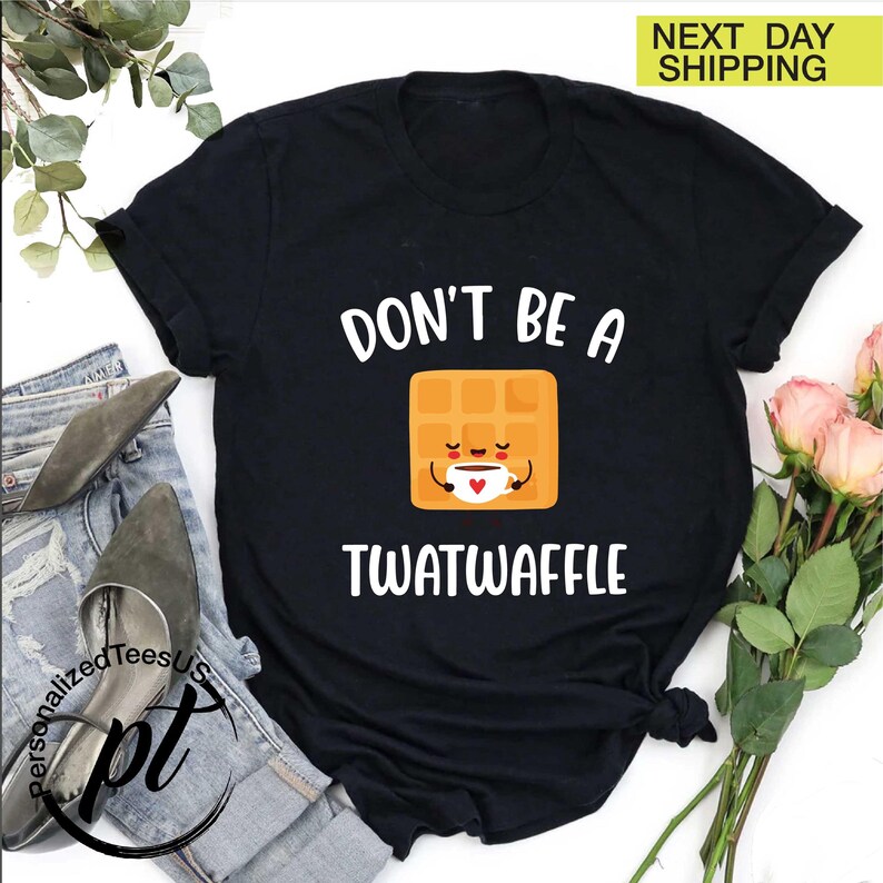 K&ouml;nnte beinhalten: Schwarzes T-Shirt mit dem wei&szlig;en Text "DON'T BE A TWATWAFFLE". Das Design zeigt eine l&auml;chelnde Waffel-Figur, die eine Kaffeetasse h&auml;lt. Das Shirt liegt auf Jeans und Schuhen.