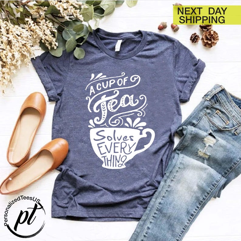 May include: A navy blue t-shirt with the text "A Cup of Tea Solves Everything" in white script. The image includes a pair of brown flats and a pair of blue jeans.