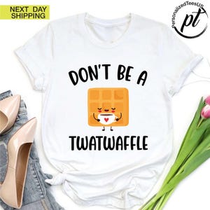 K&ouml;nnte beinhalten: Wei&szlig;es T-Shirt mit dem schwarzen Text "DON'T BE A TWATWAFFLE". Das Shirt zeigt eine Cartoon-Waffel, die eine Kaffeetasse mit einem Herzen h&auml;lt. Das Shirt hat einen Rundhalsausschnitt und kurze &Auml;rmel.