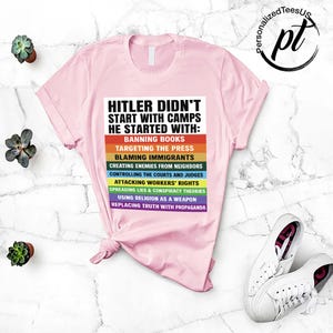 May include: A light pink t-shirt with the text "HITLER DIDN'T START WITH CAMPS HE STARTED WITH:" followed by a list of actions in a rainbow color scheme. White sneakers are visible in the bottom right corner.