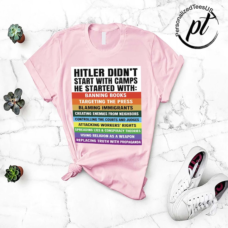 May include: A light pink t-shirt with the text "HITLER DIDN'T START WITH CAMPS HE STARTED WITH:" followed by a list of actions in a rainbow color scheme. White sneakers are visible in the bottom right corner.