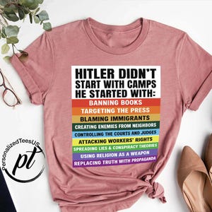 May include: A dusty rose-colored t-shirt with the text "HITLER DIDN'T START WITH CAMPS HE STARTED WITH:" followed by a list of actions in various colors. The shirt has short sleeves and a crew neck.