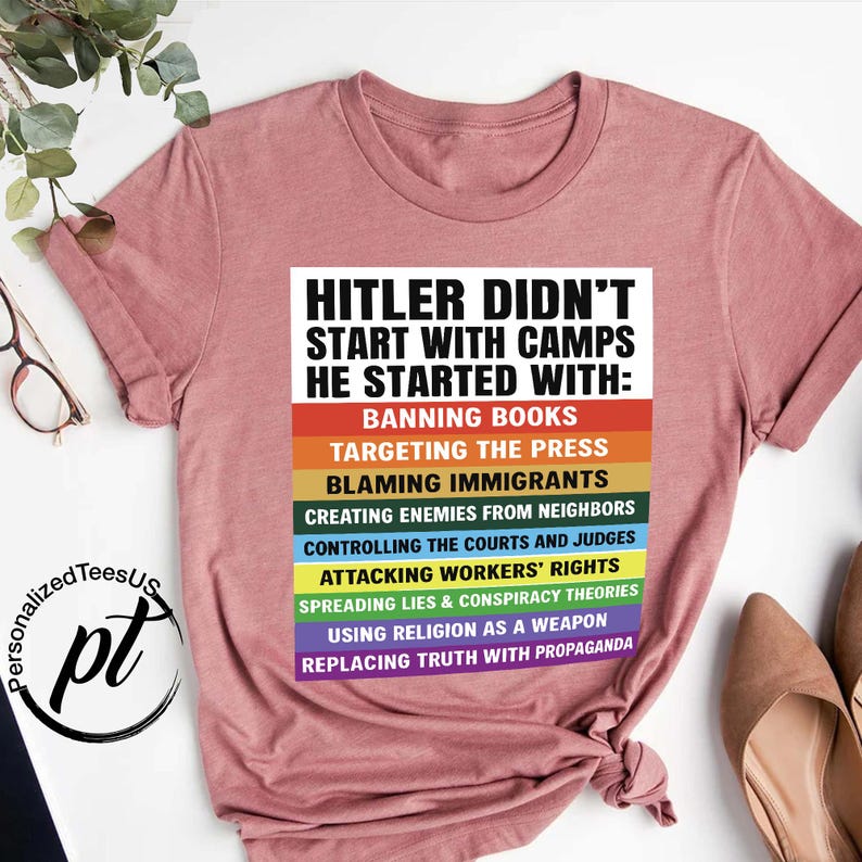 May include: A dusty rose-colored t-shirt with the text "HITLER DIDN'T START WITH CAMPS HE STARTED WITH:" followed by a list of actions in various colors. The shirt has short sleeves and a crew neck.