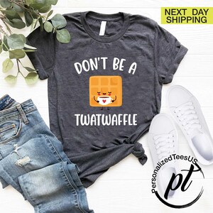 K&ouml;nnte beinhalten: Dunkelgraues T-Shirt mit dem Text "DON'T BE A TWATWAFFLE" und einer Cartoon-Waffel, die eine Kaffeetasse h&auml;lt. Das Shirt wird mit blauen Jeans und wei&szlig;en Turnschuhen kombiniert.