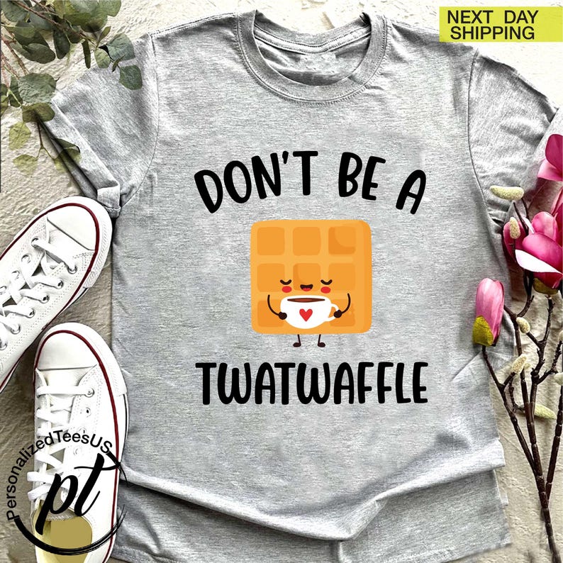K&ouml;nnte beinhalten: Hellgraues T-Shirt mit dem schwarzen Schriftzug "DON'T BE A TWATWAFFLE". In der Mitte befindet sich eine Cartoon-Waffel, die eine Kaffeetasse h&auml;lt. Wei&szlig;e Turnschuhe sind links. "NEXT DAY SHIPPING" ist oben rechts.