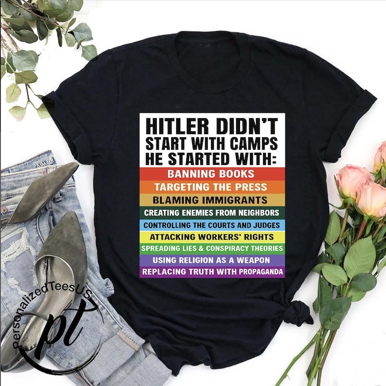 May include: Black t-shirt with the text "HITLER DIDN'T START WITH CAMPS HE STARTED WITH:" followed by a list of actions in rainbow-colored blocks. The shirt is displayed on a pair of jeans with a pair of shoes and a bouquet of flowers.