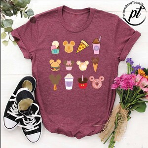 May include: A maroon t-shirt with a variety of Disney-themed food items printed on it. The food items include Mickey Mouse-shaped ice cream, pizza, popcorn, and a donut.