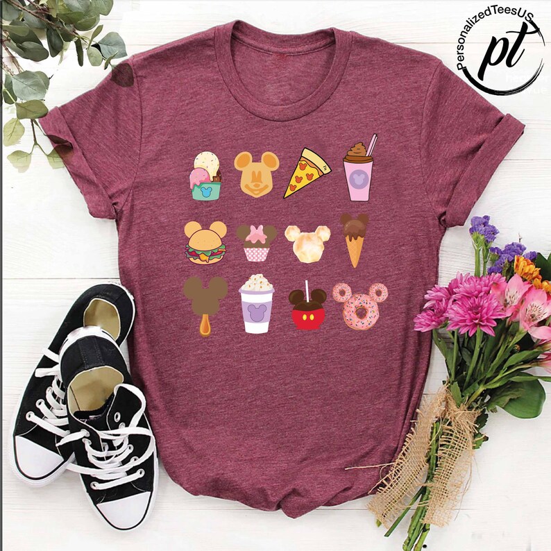 May include: A maroon t-shirt with a variety of Disney-themed food items printed on it. The food items include Mickey Mouse-shaped ice cream, pizza, popcorn, and a donut.