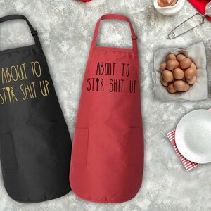 May include: Two aprons, one black and one red, with the text "About to Stir Shit Up" printed on them. The aprons have a pocket on the front.