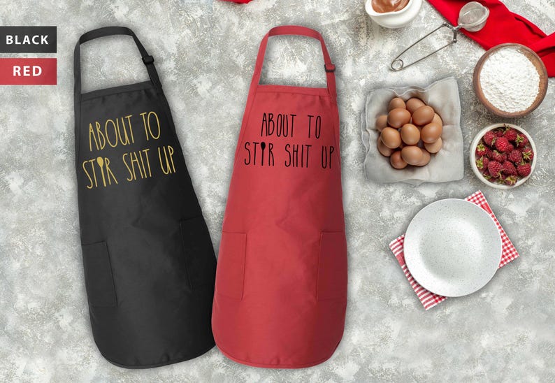May include: Two aprons, one black and one red, with the text "About to Stir Shit Up" printed on them. The aprons have a pocket on the front.