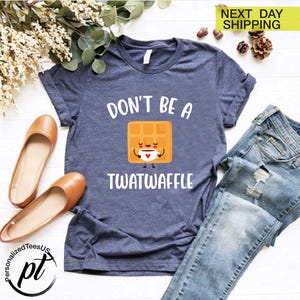 K&ouml;nnte beinhalten: Ein marineblaues T-Shirt mit dem Text "DON'T BE A TWATWAFFLE" und einer Cartoon-Waffel, die eine Kaffeetasse h&auml;lt. Das Shirt wird mit einer blauen Jeans und braunen Ballerinas kombiniert. Die Worte "NEXT DAY SHIPPING" stehen oben rechts.