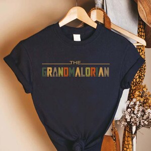 Puede incluir: Camiseta azul marino con el texto "THE GRANDMALORIAN" en una fuente de estilo retro. El texto es multicolor, con verde, naranja y marr&oacute;n. La camiseta est&aacute; colgada en una percha de madera.