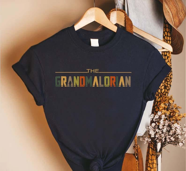 Puede incluir: Camiseta azul marino con el texto "THE GRANDMALORIAN" en una fuente de estilo retro. El texto es multicolor, con verde, naranja y marr&oacute;n. La camiseta est&aacute; colgada en una percha de madera.