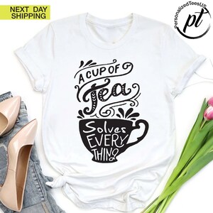 May include: White t-shirt with a black graphic that reads "A Cup of Tea Solves Everything" in a decorative font. The design features a teacup shape. The shirt is a casual wear item.