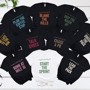 May include: A collection of nine short-sleeved t-shirts, eight in black and one in white, each featuring the text "most likely to" followed by phrases like "PLAN THE COFFEE STOP," "BLAME THE HILLS," "SKIP THE WARM UP," "BE LATE TO THE RUN," "TALK SHOES," "CHASE A PB," "CHECK THE PACE," "BONK AT MILE 10," and "START THE SPRINT."