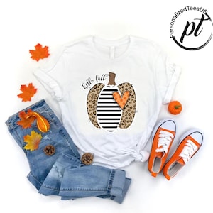 Peut inclure: T-shirt blanc avec un motif de citrouille avec des imprim&eacute;s l&eacute;opard, des rayures noires et blanches et un c&oelig;ur orange. Les mots "hello fall" sont &eacute;crits en &eacute;criture cursive. L'image comprend un jean bleu, des baskets oranges et des d&eacute;corations d'automne.