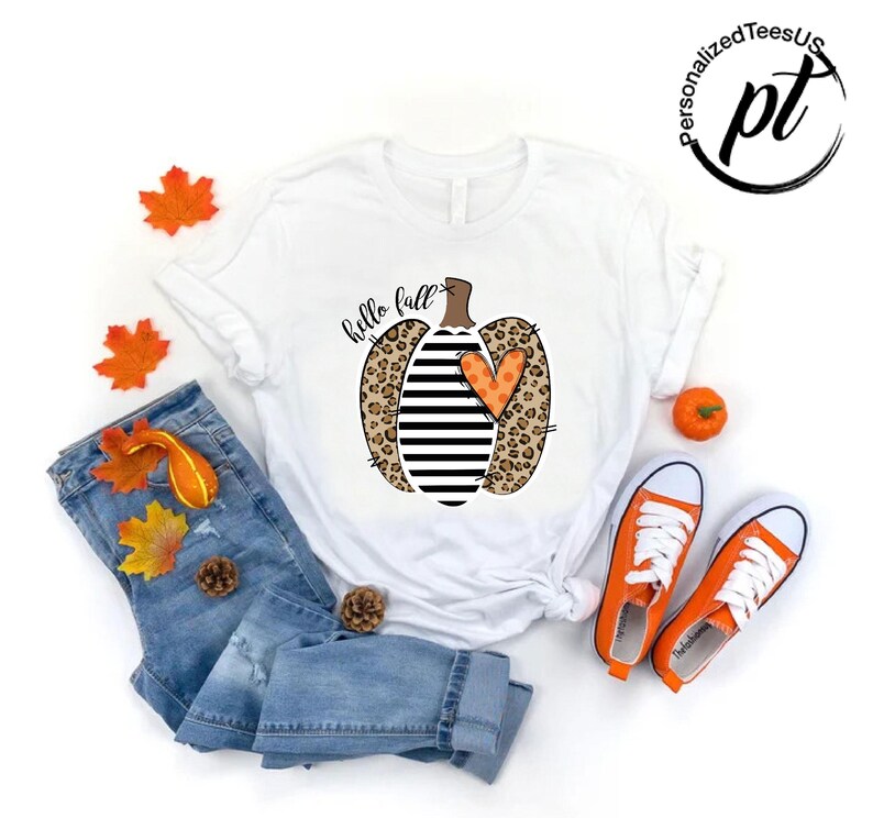 Peut inclure: T-shirt blanc avec un motif de citrouille avec des imprim&eacute;s l&eacute;opard, des rayures noires et blanches et un c&oelig;ur orange. Les mots "hello fall" sont &eacute;crits en &eacute;criture cursive. L'image comprend un jean bleu, des baskets oranges et des d&eacute;corations d'automne.