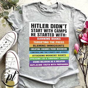 May include: A gray t-shirt with the text "HITLER DIDN'T START WITH CAMPS HE STARTED WITH:" followed by a list of actions in a rainbow-colored design. White canvas sneakers are visible on the left.