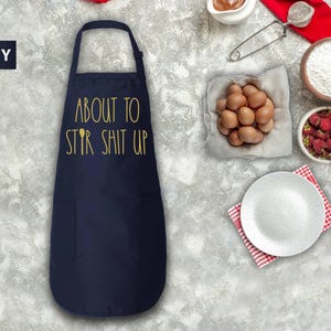 May include: A navy blue apron with the text "About to Stir Shit Up" in gold lettering. The apron has a pocket on the front.