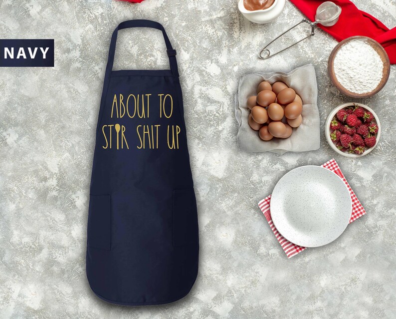 May include: A navy blue apron with the text "About to Stir Shit Up" in gold lettering. The apron has a pocket on the front.