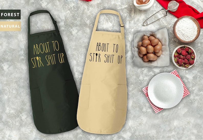 May include: Two black and beige aprons with the text "About to Stir Shit Up" printed on them. The aprons have adjustable neck straps and a pocket on the front.
