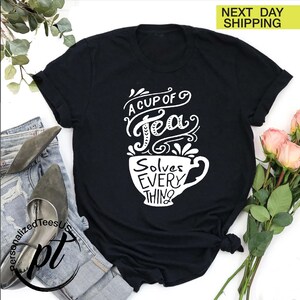 May include: Black t-shirt with a white graphic of a teacup and the words "A Cup of Tea Solves Everything". The shirt is folded, with a pair of jeans and shoes visible in the background. A bouquet of pink roses is also present.