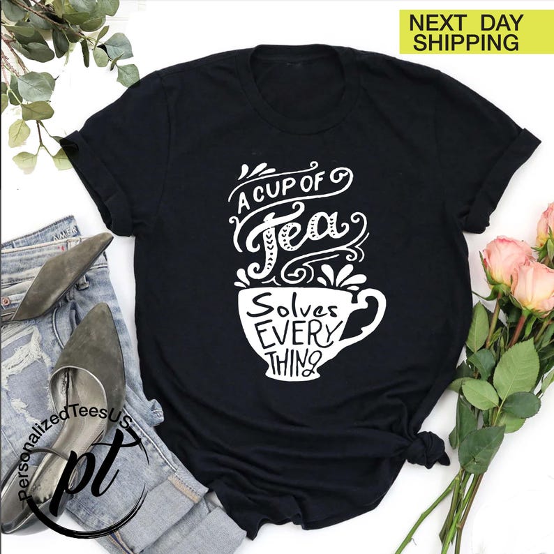 May include: Black t-shirt with a white graphic of a teacup and the words "A Cup of Tea Solves Everything". The shirt is folded, with a pair of jeans and shoes visible in the background. A bouquet of pink roses is also present.
