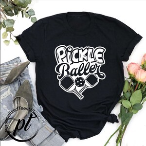 以下が含まれることがあります： 白文字で「Pickle Baller」と書かれた黒いTシャツ。その下に、白いピックルボールのパドルとボールが2つ描かれています。Tシャツは折りたたまれ、ジーンズと靴の上に置かれています。