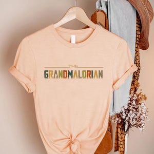 Puede incluir: Una camiseta color melocot&oacute;n con las mangas enrolladas. La camiseta presenta el texto "THE GRANDMALORIAN" en una fuente retro multicolor. La camiseta est&aacute; colgada en una percha de madera.