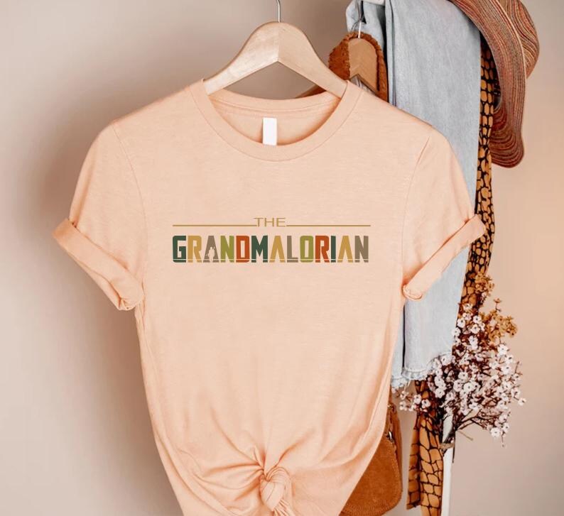 Puede incluir: Una camiseta color melocot&oacute;n con las mangas enrolladas. La camiseta presenta el texto "THE GRANDMALORIAN" en una fuente retro multicolor. La camiseta est&aacute; colgada en una percha de madera.