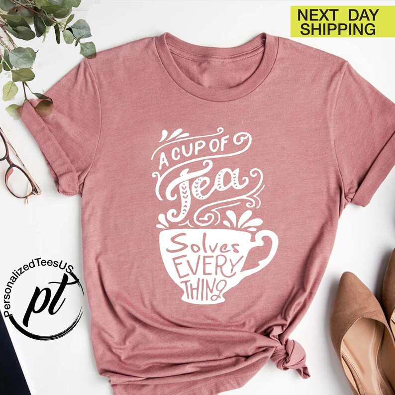 May include: A dusty rose t-shirt with the text "A Cup of Tea Solves Everything" in white script and a teacup design. The shirt has rolled-up sleeves and a crew neck. The image also includes a logo and the words "Next Day Shipping".