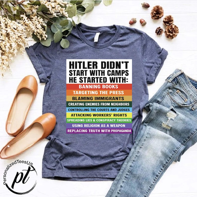 May include: A navy blue t-shirt with the text "HITLER DIDN'T START WITH CAMPS HE STARTED WITH:" and a list of actions in various colors. The shirt is paired with blue jeans and brown flats. The image includes the logo "PersonalizedTeesUS".