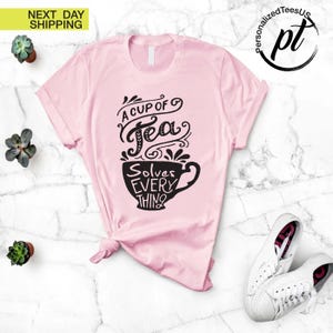 May include: A light pink t-shirt with the text "A Cup of Tea Solves Everything" in black script and a teacup design. The shirt is styled with a knot at the waist and is paired with white sneakers.