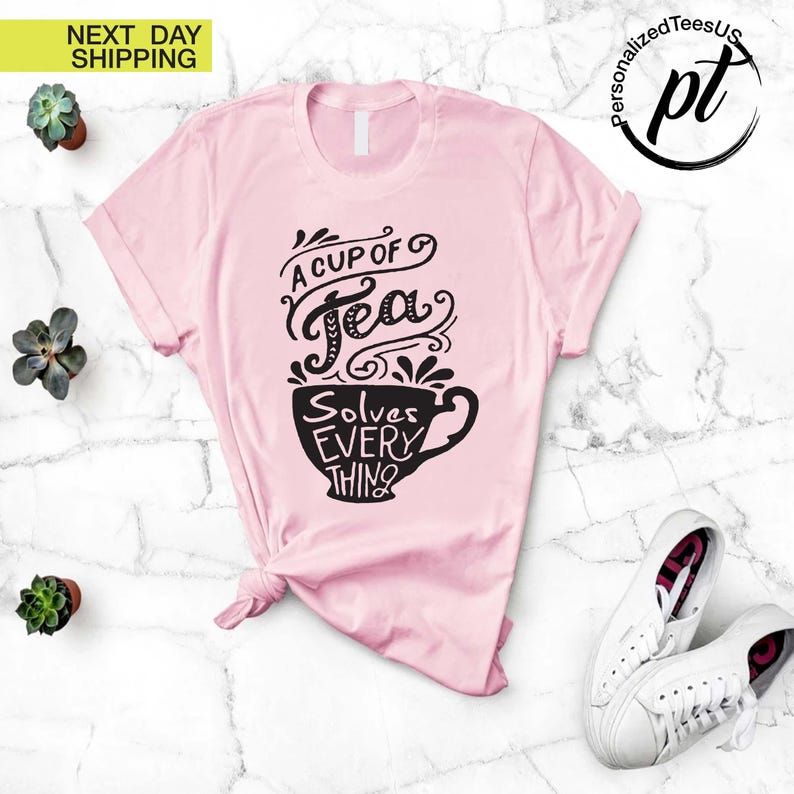 May include: A light pink t-shirt with the text "A Cup of Tea Solves Everything" in black script and a teacup design. The shirt is styled with a knot at the waist and is paired with white sneakers.