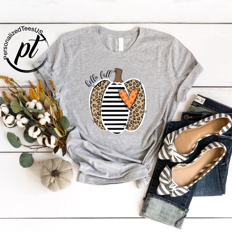 Peut inclure: T-shirt gris clair avec un motif de citrouille combinant imprim&eacute; l&eacute;opard, rayures noires et blanches et un c&oelig;ur orange. Le texte "hello fall" est au-dessus de la citrouille. Le t-shirt est associ&eacute; &agrave; un jean bleu et des ballerines ray&eacute;es.