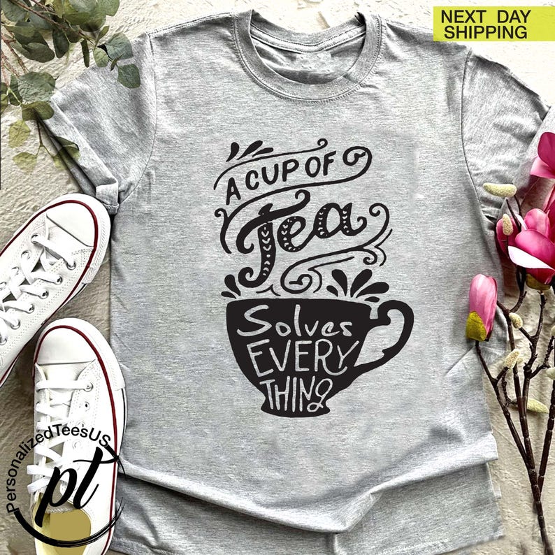 May include: A light gray t-shirt with a black graphic that reads "A Cup of Tea Solves Everything." The design features stylized lettering and a teacup illustration. White sneakers are visible on the left side of the image.