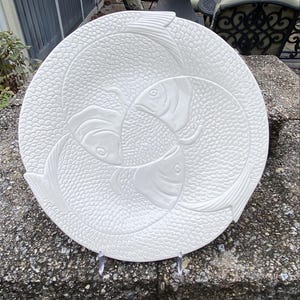 May include: A white ceramic serving platter with a raised design of three fish swimming in a circle. The fish have detailed scales and fins. The platter is round and sits on a clear stand.
